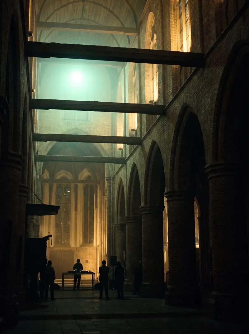 Interieur van een historische bakstenen kerk bij nacht; in de verte een kleine DJ-opstelling.
