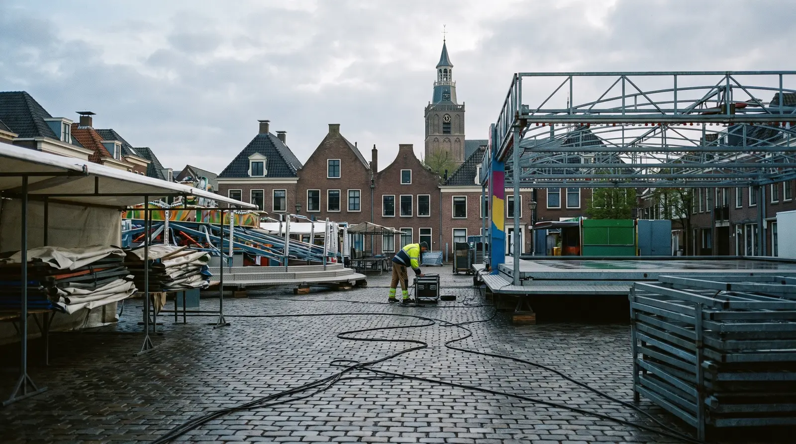 Een plein in de vroege ochtend terwijl de kermis wordt opgebouwd: stalen constructies, kabels op de keien, een kerktoren op de achtergrond.